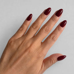 A close-up of a hand with manicured nails painted in a deep burgundy red polish, set against a plain light gray background.