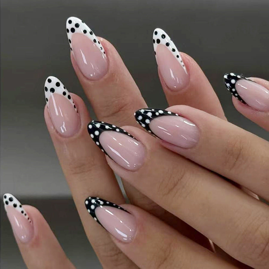 Glossy nude almond press-on nails with black and white polka dot French tip nail art on a hand model.