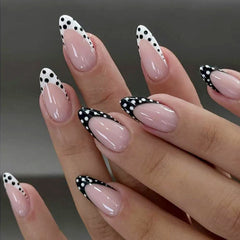 Glossy nude almond press-on nails with black and white polka dot French tip nail art on a hand model.